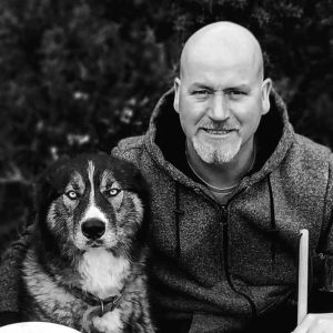 dennis-daly A black and white portrait of Dennis Daly sitting outdoors with his fluffy, wolf-like husky dog