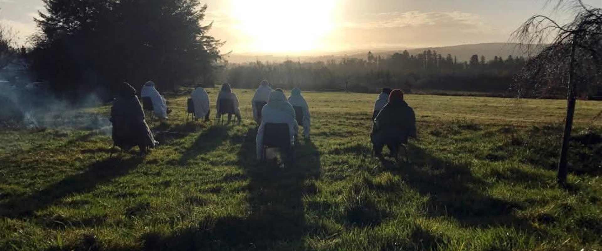 A wide panoramic view of a nature healing circle gathering, with participants seated outdoors in a quiet green field at golden hour sunrise