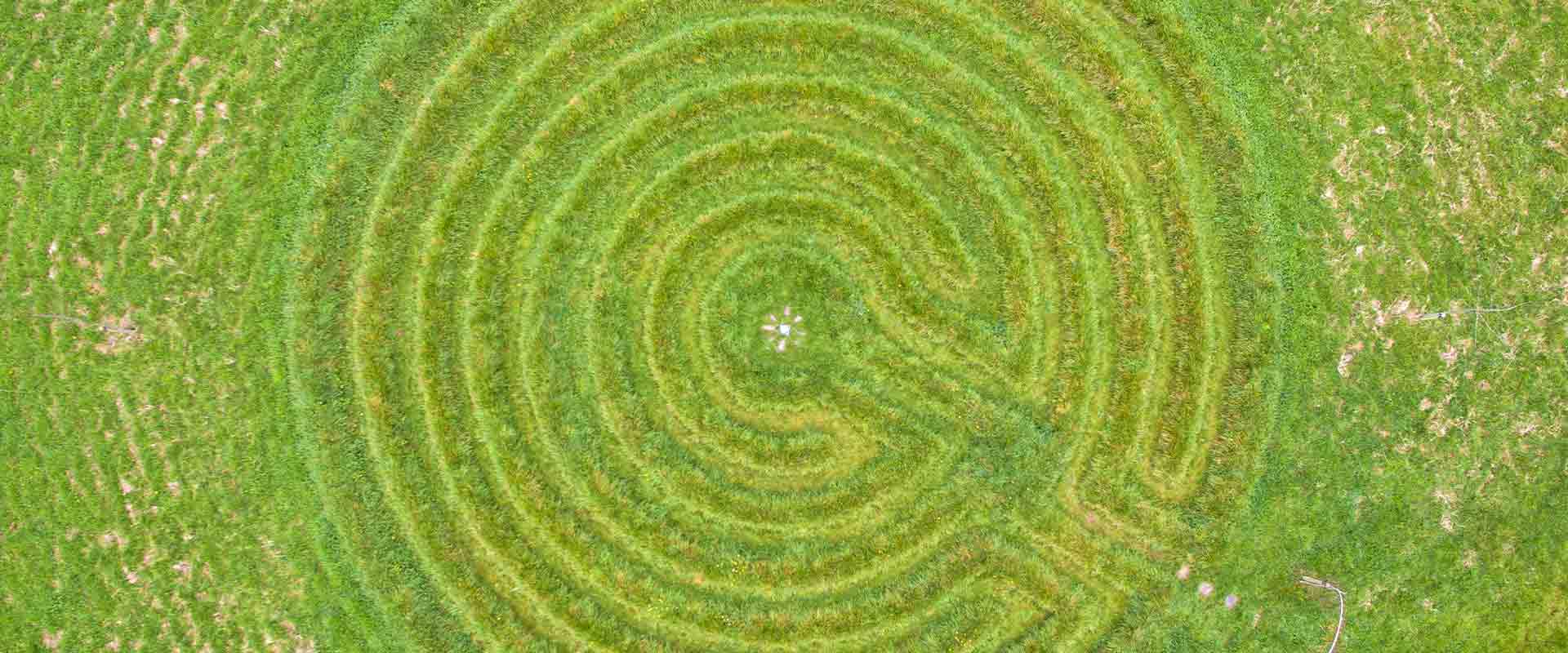 A wide panoramic, top-down drone view capturing the intricate concentric patterns of the sacred geometry grass labyrinth in Ireland